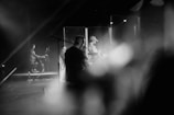 A black and white image capturing a band performing on stage with musicians playing various instruments. There is a keyboard player on one side, while other band members are playing guitar and drums. The atmosphere appears to be relaxed and focused, with soft lighting and blurry foreground elements adding depth.