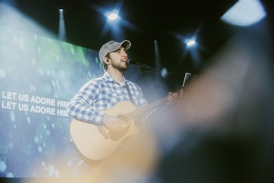 A musician wearing a checkered shirt and cap is playing an acoustic guitar and singing into a microphone on stage. Bright spotlights shine down, and a large screen in the background displays the words 'LET US ADORE HIM'. The setting appears to be a live performance or worship event.