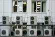 A series of air conditioning units are mounted on a metal structure outside a building with white paneling. Two large windows are visible behind the units, revealing the indoors with fluorescent lighting.