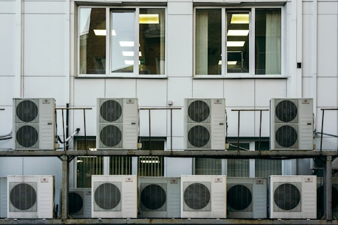 A series of air conditioning units are mounted on a metal structure outside a building with white paneling. Two large windows are visible behind the units, revealing the indoors with fluorescent lighting.