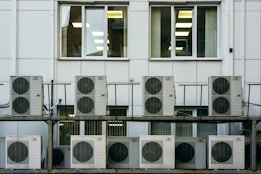 A series of air conditioning units are mounted on a metal structure outside a building with white paneling. Two large windows are visible behind the units, revealing the indoors with fluorescent lighting.