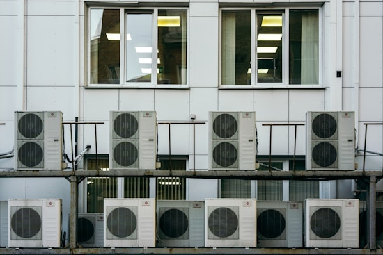 A series of air conditioning units are mounted on a metal structure outside a building with white paneling. Two large windows are visible behind the units, revealing the indoors with fluorescent lighting.