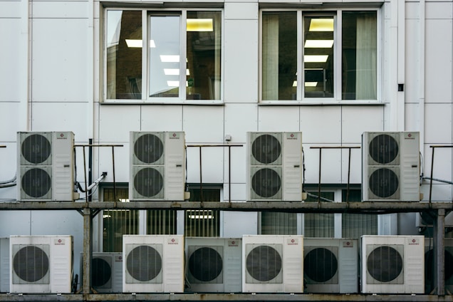 A series of air conditioning units are mounted on a metal structure outside a building with white paneling. Two large windows are visible behind the units, revealing the indoors with fluorescent lighting.