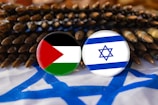 Two buttons displaying the Palestinian and Israeli flags are placed against a background of ammunition. The setting includes a fabric with the Israeli flag design, creating a contrast between symbols of national identity and conflict.