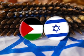 Two buttons displaying the Palestinian and Israeli flags are placed against a background of ammunition. The setting includes a fabric with the Israeli flag design, creating a contrast between symbols of national identity and conflict.