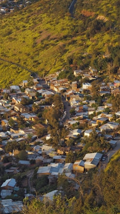 A small hillside village densely packed with houses, featuring corrugated metal roofs and narrow streets. The surrounding landscape is lush and green with a winding road leading into the community.