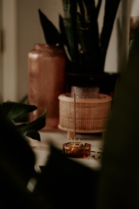 A serene and dimly lit setting featuring a burning incense stick placed in a small amber-colored holder. Surrounding the incense are various pottery and woven containers, along with lush green plants that add a natural element to the composition.