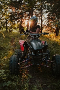 A person wearing a helmet and a black shirt sits on an all-terrain vehicle (ATV) in a forested area. The sun is setting behind the trees, casting a warm glow through the foliage. The ATV is black and red, and the ground is covered with grass and leaves.