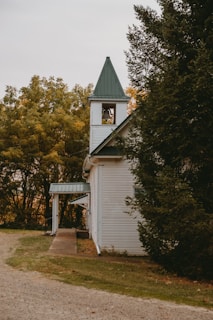 A quaint old church building is nestled among trees with a bell tower that features a green roof. The surrounding foliage shows signs of early autumn, displaying a mix of green and yellow leaves. The architecture is traditional, with white wooden siding and green roof shingles, reflecting a rustic charm.