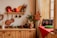 A cozy kitchen setting with wooden cabinets and a shelf displaying an assortment of copper kitchenware and red candles. Festive decorations include a small decorated Christmas tree, a vibrant star ornament, and red berries. A wooden table is adorned with evergreen branches and a red tablecloth adds a pop of color to the corner by the window.