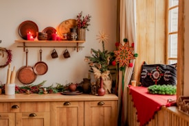 A cozy kitchen setting with wooden cabinets and a shelf displaying an assortment of copper kitchenware and red candles. Festive decorations include a small decorated Christmas tree, a vibrant star ornament, and red berries. A wooden table is adorned with evergreen branches and a red tablecloth adds a pop of color to the corner by the window.