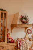 A cozy kitchen setting featuring a wooden cabinet and countertop. Decorative wreaths with red berries and green foliage adorn the walls. There is a collection of wooden cutting boards and kitchen utensils, with red mugs and a fresh loaf of bread placed on a patterned cloth. A warm, inviting ambiance is emphasized by rustic elements and natural materials.