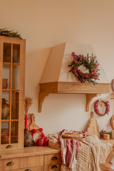 A cozy kitchen setting featuring a wooden cabinet and countertop. Decorative wreaths with red berries and green foliage adorn the walls. There is a collection of wooden cutting boards and kitchen utensils, with red mugs and a fresh loaf of bread placed on a patterned cloth. A warm, inviting ambiance is emphasized by rustic elements and natural materials.