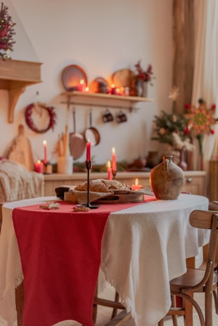 A rustic kitchen setting with a table adorned with a white cloth and a red runner, featuring a loaf of bread, a ceramic jug, and lit red candles. The background includes shelves with kitchenware and festive decor like wreaths and red berries, evoking a cozy atmosphere.