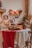 A rustic kitchen setting with a table adorned with a white cloth and a red runner, featuring a loaf of bread, a ceramic jug, and lit red candles. The background includes shelves with kitchenware and festive decor like wreaths and red berries, evoking a cozy atmosphere.