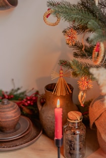 A cozy and rustic scene with a decorated Christmas tree featuring straw and wooden ornaments. A lit red candle stands in the foreground, casting a warm glow. An earthenware jug and plates, along with a glass jar filled with small natural objects, are positioned on a wooden surface.