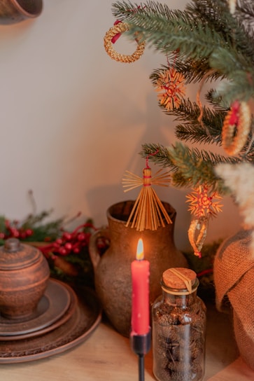 A cozy and rustic scene with a decorated Christmas tree featuring straw and wooden ornaments. A lit red candle stands in the foreground, casting a warm glow. An earthenware jug and plates, along with a glass jar filled with small natural objects, are positioned on a wooden surface.
