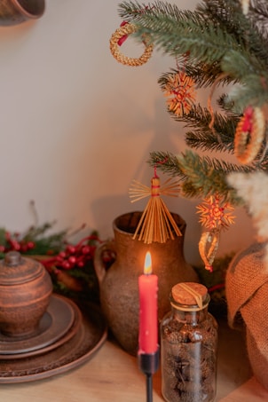 A cozy and rustic scene with a decorated Christmas tree featuring straw and wooden ornaments. A lit red candle stands in the foreground, casting a warm glow. An earthenware jug and plates, along with a glass jar filled with small natural objects, are positioned on a wooden surface.