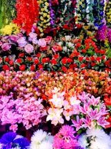 A vibrant and colorful collection of various artificial flowers arranged densely in layers. The display features a wide range of colors, including red, pink, yellow, white, purple, and greens. The flowers are a mix of roses, lilies, daisies, and other exotic blooms, creating a rich and lush visual appeal.