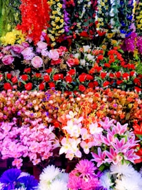 A vibrant and colorful collection of various artificial flowers arranged densely in layers. The display features a wide range of colors, including red, pink, yellow, white, purple, and greens. The flowers are a mix of roses, lilies, daisies, and other exotic blooms, creating a rich and lush visual appeal.