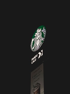 A Starbucks drive-thru sign is illuminated against a dark background. The iconic Starbucks logo is prominently displayed, with an arrow indicating the direction for the drive-thru. Below, a poster advertises a frappuccino with text.