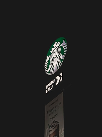 A Starbucks drive-thru sign is illuminated against a dark background. The iconic Starbucks logo is prominently displayed, with an arrow indicating the direction for the drive-thru. Below, a poster advertises a frappuccino with text.
