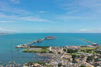 An aerial view of a coastal area with a large body of turquoise water extending into the distance. A port or harbor is visible, with ships and boats scattered throughout the water. The land features buildings, some industrial structures, and a densely populated urban area near the waterfront. A long pier or jetty extends into the sea, creating a sheltered area for vessels.