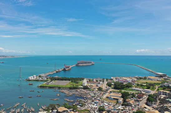 An aerial view of a coastal area with a large body of turquoise water extending into the distance. A port or harbor is visible, with ships and boats scattered throughout the water. The land features buildings, some industrial structures, and a densely populated urban area near the waterfront. A long pier or jetty extends into the sea, creating a sheltered area for vessels.