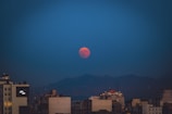 A blood-red full moon is prominently visible against a dark blue sky, partially above the horizon. Below, a city skyline features various buildings that are silhouetted against the mountainous backdrop.
