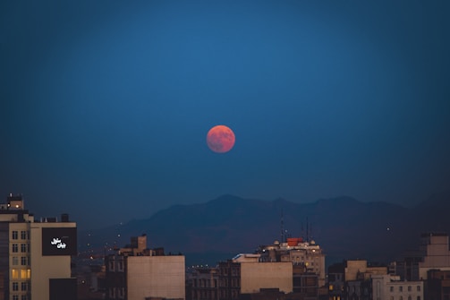 A blood-red full moon is prominently visible against a dark blue sky, partially above the horizon. Below, a city skyline features various buildings that are silhouetted against the mountainous backdrop.