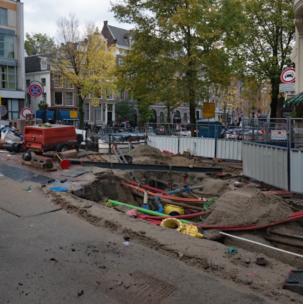 A city street under construction, with a large trench filled with colorful pipes and sand. A red generator and traffic cones are nearby, surrounded by temporary metal fencing. Trees and buildings line the street, with signs indicating road closures.