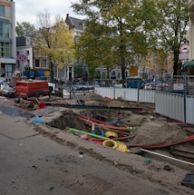 A city street under construction, with a large trench filled with colorful pipes and sand. A red generator and traffic cones are nearby, surrounded by temporary metal fencing. Trees and buildings line the street, with signs indicating road closures.