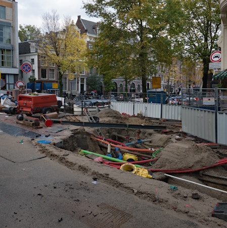 A city street under construction, with a large trench filled with colorful pipes and sand. A red generator and traffic cones are nearby, surrounded by temporary metal fencing. Trees and buildings line the street, with signs indicating road closures.
