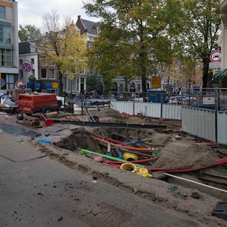 A city street under construction, with a large trench filled with colorful pipes and sand. A red generator and traffic cones are nearby, surrounded by temporary metal fencing. Trees and buildings line the street, with signs indicating road closures.