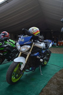 Two motorcycles are parked on a green carpet under a large tent. The motorcycle in the foreground is primarily blue with neon green wheels and a visible license plate. The rider is wearing a helmet with cartoon graphics. The second motorcycle, partially visible, is black and green. There are people and chairs in the background, suggesting a gathering or event.