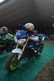 Two motorcycles are parked on a green carpet under a large tent. The motorcycle in the foreground is primarily blue with neon green wheels and a visible license plate. The rider is wearing a helmet with cartoon graphics. The second motorcycle, partially visible, is black and green. There are people and chairs in the background, suggesting a gathering or event.