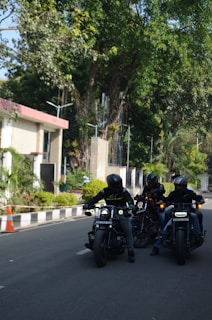 Three motorcyclists wearing helmets are riding their bikes on a road bordered by greenery and buildings. The scene is set in a lush, leafy environment with tall trees and small bushes lining the road. One of the buildings has a red roof and modern architecture. The riders are grouped closely together with their headlights on, creating a sense of camaraderie.