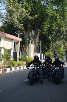 Three motorcyclists wearing helmets are riding their bikes on a road bordered by greenery and buildings. The scene is set in a lush, leafy environment with tall trees and small bushes lining the road. One of the buildings has a red roof and modern architecture. The riders are grouped closely together with their headlights on, creating a sense of camaraderie.