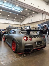 A sleek, customized sports car with a matte gray finish and red accents is displayed in a large indoor venue. The car features a prominent rear spoiler and aftermarket modifications, suggesting it is showcased at an automotive exhibition. Bright overhead lights and banners can be seen in the spacious hall.