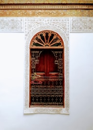 A richly decorated wooden door is framed by intricate white lace-like carvings against a white wall. The upper part of the frame has an arch with detailed, filigree patterns. There is a metal railing with hanging clothes hangers in front of the door.