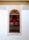 A richly decorated wooden door is framed by intricate white lace-like carvings against a white wall. The upper part of the frame has an arch with detailed, filigree patterns. There is a metal railing with hanging clothes hangers in front of the door.