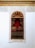 A richly decorated wooden door is framed by intricate white lace-like carvings against a white wall. The upper part of the frame has an arch with detailed, filigree patterns. There is a metal railing with hanging clothes hangers in front of the door.