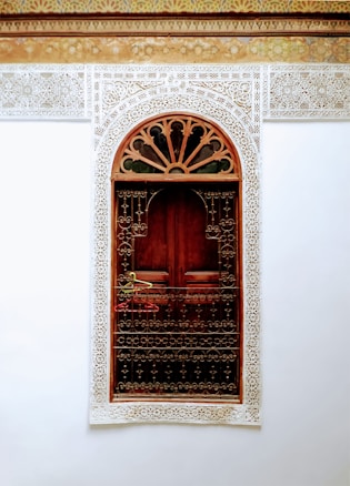 A richly decorated wooden door is framed by intricate white lace-like carvings against a white wall. The upper part of the frame has an arch with detailed, filigree patterns. There is a metal railing with hanging clothes hangers in front of the door.