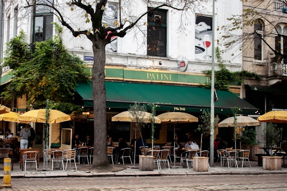 A street-side café features a green canopy with yellow umbrellas, metal tables, and chairs. The building is covered with vines and has a tree growing in front. Patrons are seated under the umbrellas, with a sign displaying 'PATINE'. The setting appears inviting, with a mix of natural and urban elements.