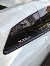 A close-up view of a car window featuring a decal that reads 'GT Owners Club R Indonesia' with a reflection of a cityscape visible on the window. The door and window frame are visible, with greenery and a parked car in the background.