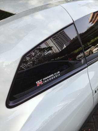 A close-up view of a car window featuring a decal that reads 'GT Owners Club R Indonesia' with a reflection of a cityscape visible on the window. The door and window frame are visible, with greenery and a parked car in the background.