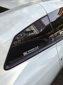 A close-up view of a car window featuring a decal that reads 'GT Owners Club R Indonesia' with a reflection of a cityscape visible on the window. The door and window frame are visible, with greenery and a parked car in the background.