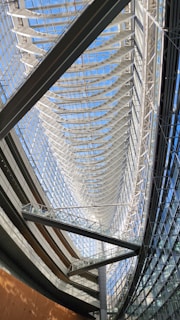 A spacious and modern interior of a building features a large glass ceiling structure allowing natural light to flood in. The design incorporates sleek curved elements with a series of parallel lines and beams, creating a sense of movement and openness. Walkways and terraces add to the architectural complexity.