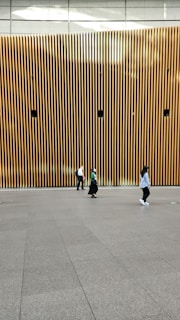 A modern architectural wall featuring vertical wooden slats with several circular speakers or installations evenly spaced. Three people are walking across a smooth, spacious floor in front of the wall, each dressed casually.