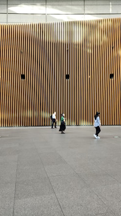 A modern architectural wall featuring vertical wooden slats with several circular speakers or installations evenly spaced. Three people are walking across a smooth, spacious floor in front of the wall, each dressed casually.